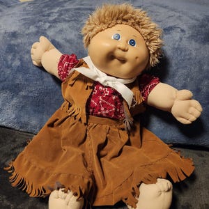 Può includere: Una bambola Cabbage Patch Kid che indossa una gonna di camoscio marrone con frange, un bandano rosso e bianco e un gilet bianco con frange. La bambola ha i capelli biondi e gli occhi azzurri.