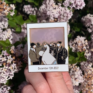 May include: A gold-toned enamel pin featuring a group of people wearing face masks, standing in a line and holding hands. The pin is in the shape of a Polaroid photo with the date "December 13th 2022" printed below the image.