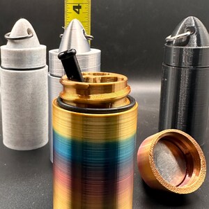 May include: A cylindrical container with a gradient finish of gold, blue, and purple, featuring a black O-ring and a black insert. A yellow measuring tape shows the height is approximately 4.5 inches. Two additional containers in gray and black are visible.