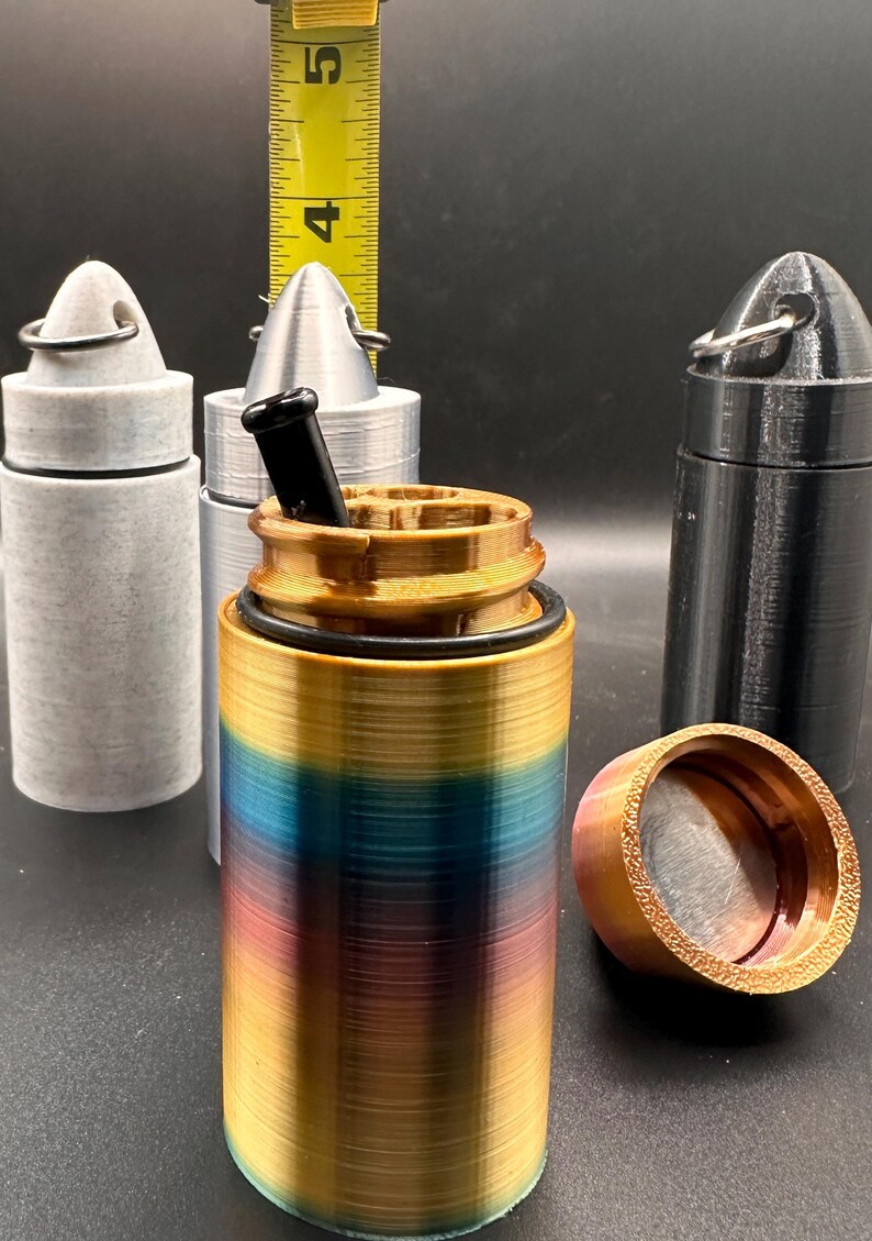 May include: A cylindrical container with a gradient finish of gold, blue, and purple, featuring a black O-ring and a black insert. A yellow measuring tape shows the height is approximately 4.5 inches. Two additional containers in gray and black are visible.