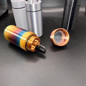 May include: A collection of cylindrical containers with pointed caps and metal rings. One container is multicolored with a gradient of yellow, blue, and red, featuring an open top with a black nozzle. Two other containers are solid gray and black.