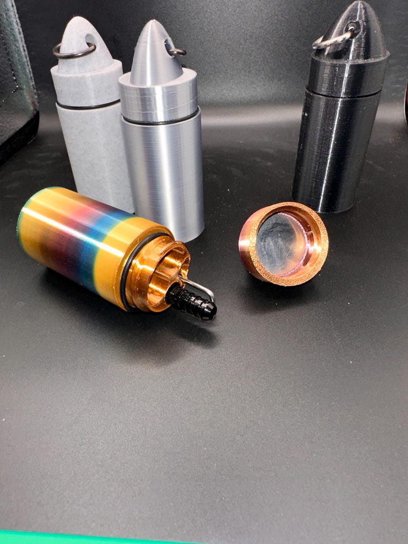 May include: A collection of cylindrical containers with pointed caps and metal rings. One container is multicolored with a gradient of yellow, blue, and red, featuring an open top with a black nozzle. Two other containers are solid gray and black.
