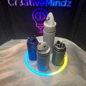 May include: Four cylindrical containers with metal key rings attached to their tops. One is blue, one is light gray, one is dark gray, and one is white. The blue and dark gray containers are open, revealing internal compartments and a small red tool.