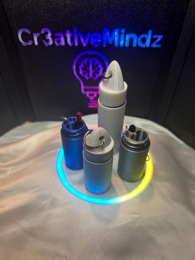 May include: Four cylindrical containers with metal key rings attached to their tops. One is blue, one is light gray, one is dark gray, and one is white. The blue and dark gray containers are open, revealing internal compartments and a small red tool.