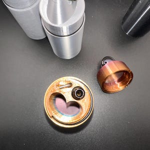 May include: A collection of cylindrical containers with pointed caps and metal rings. One copper-colored container is open, revealing a heart-shaped interior cavity and a small black nozzle. Two grey containers stand upright, and a separate copper cap sits nearby.