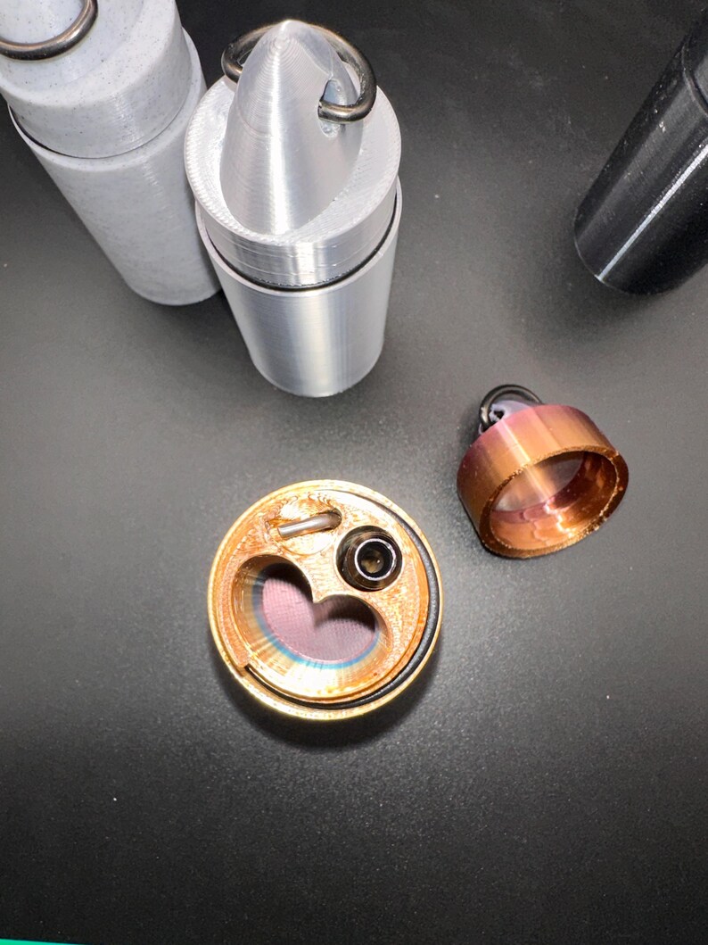 May include: A collection of cylindrical containers with pointed caps and metal rings. One copper-colored container is open, revealing a heart-shaped interior cavity and a small black nozzle. Two grey containers stand upright, and a separate copper cap sits nearby.