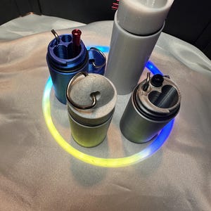 May include: A collection of four cylindrical containers with lids, including one blue, one grey, one speckled off-white, and one tall white container. Each features a metal ring attachment at the top, and the blue and grey containers have internal compartments.