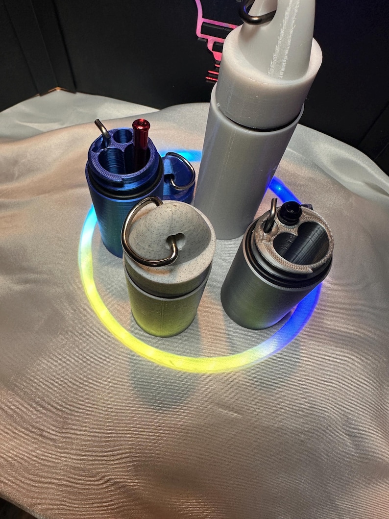 May include: A collection of four cylindrical containers with lids, including one blue, one grey, one speckled off-white, and one tall white container. Each features a metal ring attachment at the top, and the blue and grey containers have internal compartments.