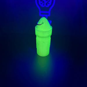 May include: A cylindrical, neon green, 3D-printed container with a rounded cap and a metal ring attachment. The object glows under ultraviolet light, positioned in front of a glowing blue light bulb sign containing a brain icon and partial text.