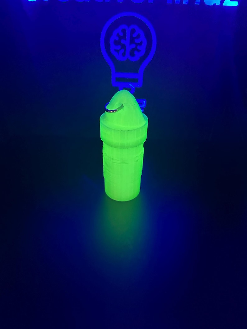 May include: A cylindrical, neon green, 3D-printed container with a rounded cap and a metal ring attachment. The object glows under ultraviolet light, positioned in front of a glowing blue light bulb sign containing a brain icon and partial text.