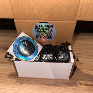May include: A small white cardboard shipping box filled with black shredded paper filler. A circular sticker with a QR code is on the inside back flap, and a square holographic sticker featuring a skull and the text Cr3ativeMindz is attached.