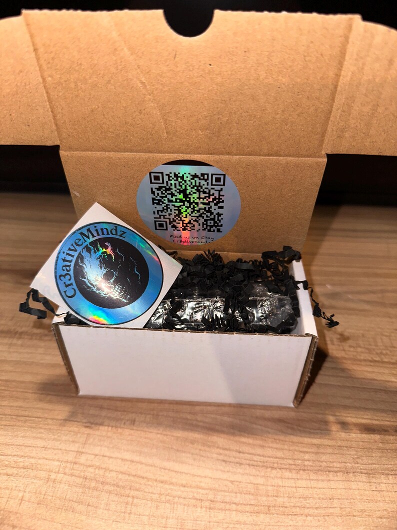 May include: A small white cardboard shipping box filled with black shredded paper filler. A circular sticker with a QR code is on the inside back flap, and a square holographic sticker featuring a skull and the text Cr3ativeMindz is attached.