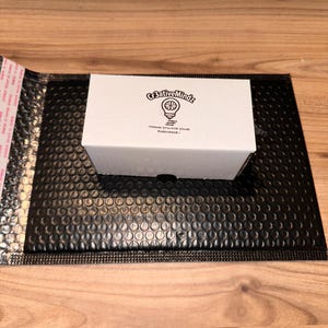 May include: A rectangular white cardboard box featuring a black printed logo of a lightbulb with the text Cr3ativeMindz and the phrase THANK YOU FOR YOUR PURCHASE centered on the top surface, resting on a black bubble mailer envelope.
