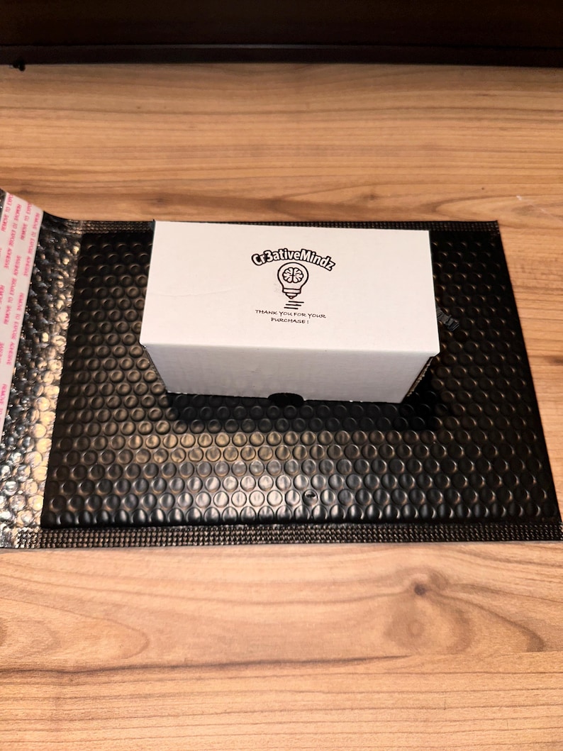 May include: A rectangular white cardboard box featuring a black printed logo of a lightbulb with the text Cr3ativeMindz and the phrase THANK YOU FOR YOUR PURCHASE centered on the top surface, resting on a black bubble mailer envelope.