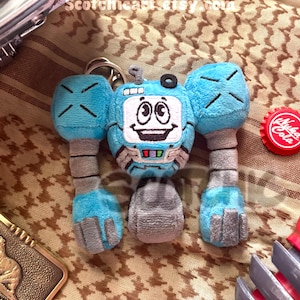 May include: A plush robot keychain in shades of blue, gray, and white. The robot has a screen face with a smiling expression. A red bottle cap with white text is visible. The text "Scotchichart.etsy.com" is visible.