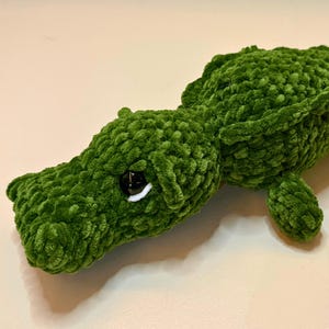 Puede incluir: Un juguete de cocodrilo verde tejido a ganchillo con ojos blancos y negros. El reptil hecho a mano tiene un aspecto texturizado y aterciopelado y está tumbado. La imagen también incluye un gráfico de patrón de ganchillo de archivo digital.