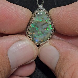 May include: A pear-shaped pendant with a silver-colored setting. The pendant features a vibrant opal with flashes of green, blue, and orange. The setting has a decorative, scalloped edge and a small loop for a chain.