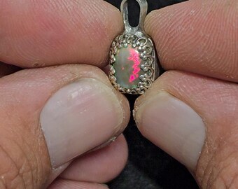May include: A small, silver pendant with an oval opal gemstone. The opal displays a play of color with pink and green hues. The pendant has a decorative, filigree-style border. The pendant is held between two fingers.