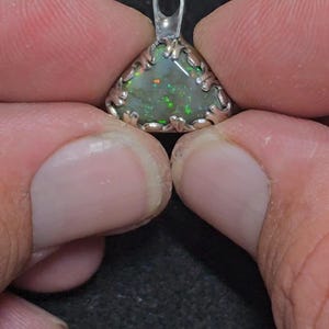 May include: A triangular pendant with a silver-colored setting. The pendant features a vibrant, iridescent opal stone with green and orange hues. The opal is set within a decorative, openwork frame, and has a small loop at the top for a chain.