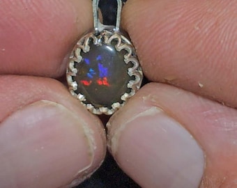 May include: A small, oval pendant with a dark gemstone displaying flashes of red and blue. The gemstone is set in a silver-colored, scalloped bezel. The pendant is held between two fingers.