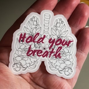 May include: A white sticker with a black outline of a pair of lungs with flowers around them. The text "Hold your breath" is written in pink inside the lungs.