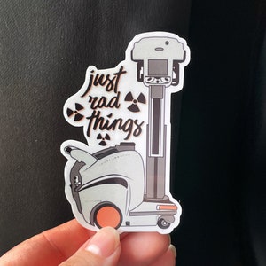 May include: A white sticker with a cartoon robot and the text "just rad things". The robot has a black and white design with orange wheels. The sticker has a black and white radioactive symbol on each side of the text.