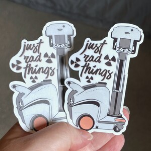 May include: Two white and gray robot stickers with the text "just rad things" and a radioactive symbol. The robots have orange wheels and are holding a small, gray object.