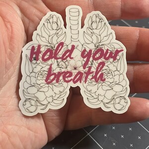 May include: A white sticker with a black outline of lungs with flowers around them. The text "Hold your breath" is written in pink inside the lungs.