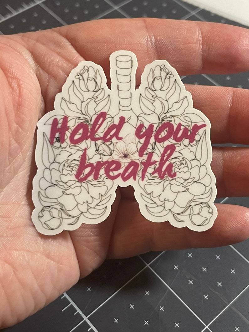 May include: A white sticker with a black outline of lungs with flowers around them. The text "Hold your breath" is written in pink inside the lungs.