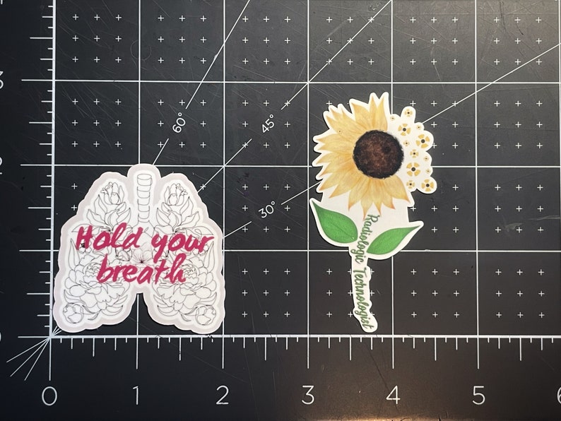 May include: Two stickers, one with a white outline of lungs with pink flowers and the text "Hold your breath", and the other with a yellow sunflower and the text "Radiologic Technologist".