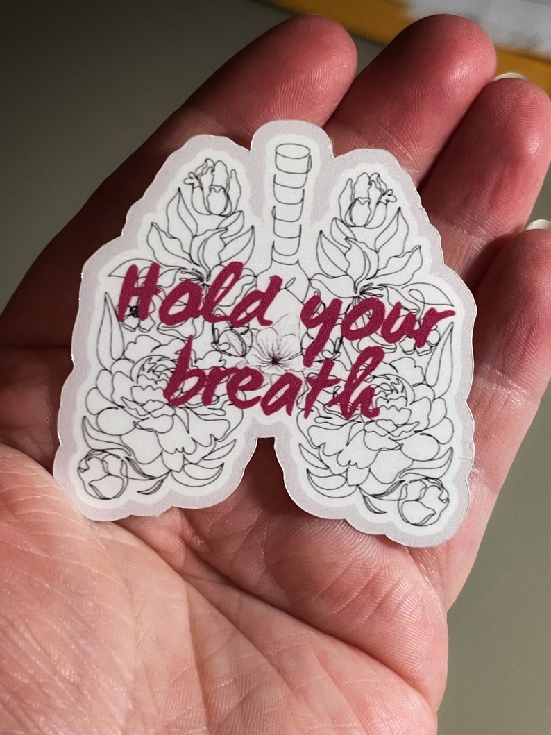 May include: A white sticker with a black outline of a pair of lungs with flowers around them. The text "Hold your breath" is written in pink inside the lungs.