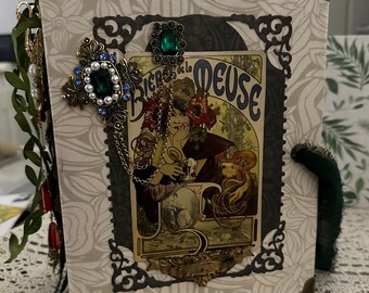 May include: A vintage-style journal with a decorative cover featuring a floral pattern, a gold-colored metal corner piece, and a black and gold ornate frame with a vintage poster inside. The poster features a woman with a green and blue jeweled headpiece and the text "Biere de la Deuse".
