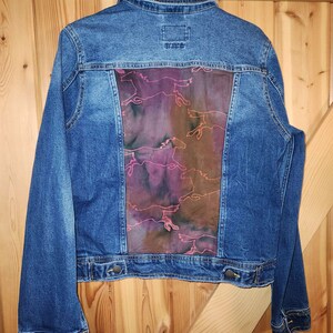 May include: Blue denim jacket with a rectangular back panel featuring a running horse design in shades of pink, purple, and brown. The jacket has button closures and a classic collar.