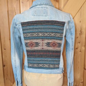 May include: A light blue denim jacket with a colorful patterned fabric patch on the back. The patch features geometric designs in shades of blue, brown, and red.