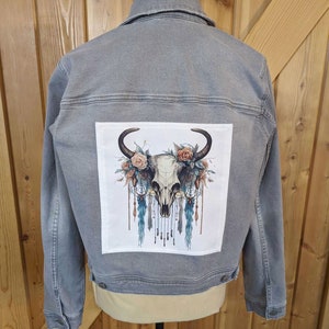 May include: A gray denim jacket with a white patch on the back featuring a skull with floral accents and dreamcatchers.