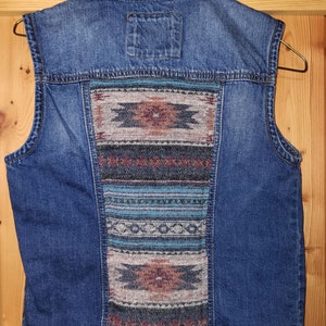May include: A blue denim vest with a colorful patterned fabric panel on the back. The panel features geometric designs in shades of brown, blue, and white.