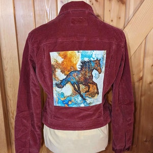 Puede incluir: Una chaqueta de pana marrón con un parche en la espalda. El parche presenta un diseño abstracto colorido con una silueta de un caballo corriendo.