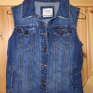 May include: A blue denim vest with a frayed hem and a button closure. The vest has two front pockets and a brand label that reads "Mudd" with the size "S" visible.