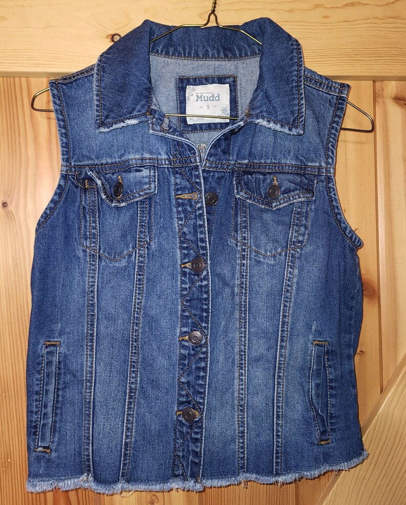May include: A blue denim vest with a frayed hem and a button closure. The vest has two front pockets and a brand label that reads "Mudd" with the size "S" visible.