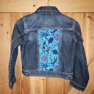 May include: A blue denim jacket with a colourful patterned fabric patch on the back. The patch features a blue background with stylised figures, turtles, and suns in shades of pink, purple, and green.