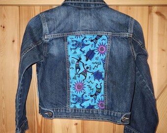 May include: A blue denim jacket with a colorful patterned fabric patch on the back. The patch features a blue background with stylized figures, turtles, and suns in shades of pink, purple, and green.