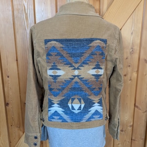 May include: Tan corduroy jacket with a geometric patterned back panel in shades of blue, brown, and cream. The jacket has a button-down front, long sleeves, and a classic collar. The back panel features a Southwestern-inspired design.