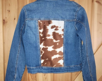 May include: A blue denim jacket with a brown and white cowhide panel on the back. The jacket has a classic collar, button closures, and long sleeves. The cowhide panel adds a unique, rustic touch to the garment.