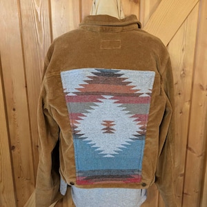 May include: A brown corduroy jacket with a Southwestern-style patterned back panel in shades of gray, blue, red, and brown. The jacket has a button closure and a collar.
