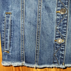 May include: Close-up of a blue denim jacket with frayed edges and two metal buttons. The jacket has a pocket on the left side and stitching details.