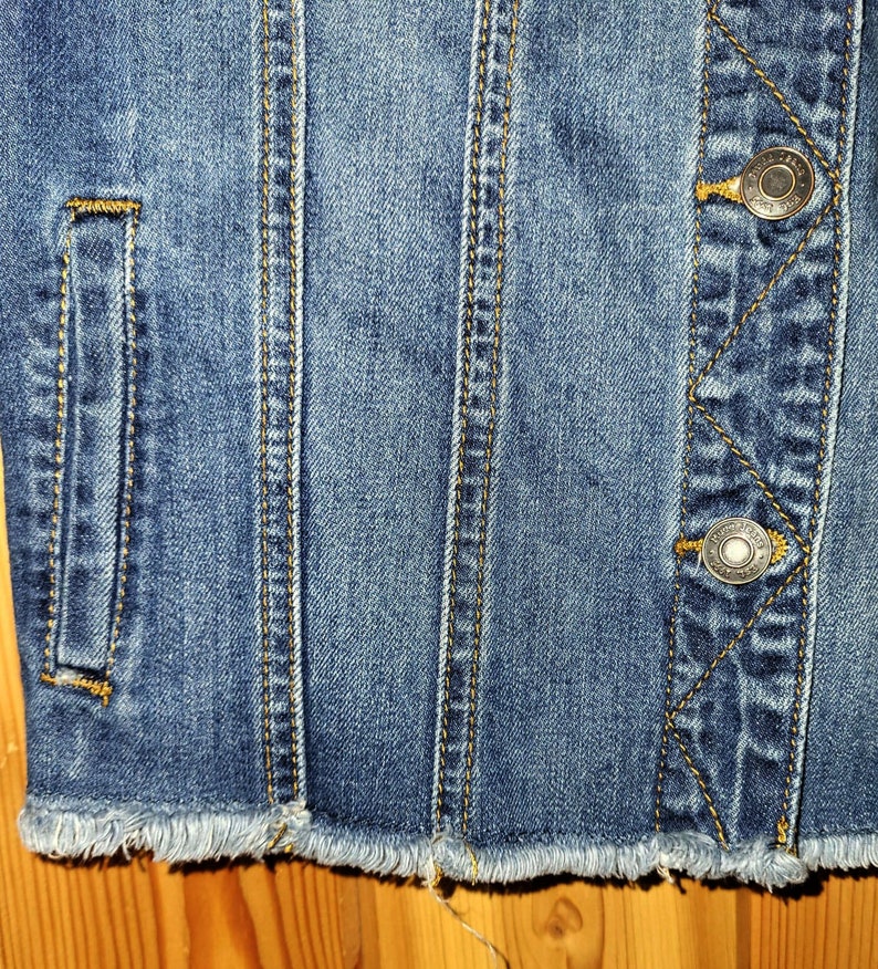 May include: Close-up of a blue denim jacket with frayed edges and two metal buttons. The jacket has a pocket on the left side and stitching details.