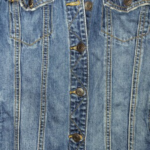 May include: Close-up of a blue denim jacket with a button closure and a pocket on the right side. The jacket has a distressed look with frayed edges.