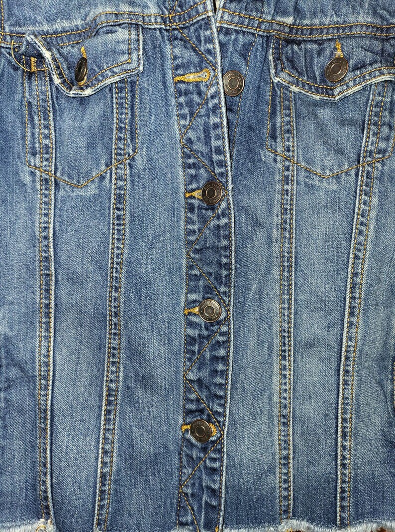 May include: Close-up of a blue denim jacket with a button closure and a pocket on the right side. The jacket has a distressed look with frayed edges.