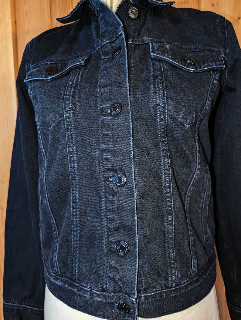 May include: A dark blue denim jacket with a classic style. The jacket has a button-up front, two chest pockets, and long sleeves.