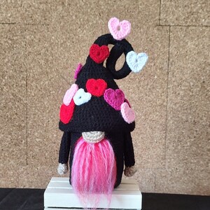 Puede incluir: Un gnomo de San Valentín hecho a mano con un sombrero negro adornado con corazones rojos, rosas y blancos. El gnomo tiene una barba rosa y se exhibe sobre una caja de madera blanca. Corazones de ganchillo adicionales están dispersos al frente.